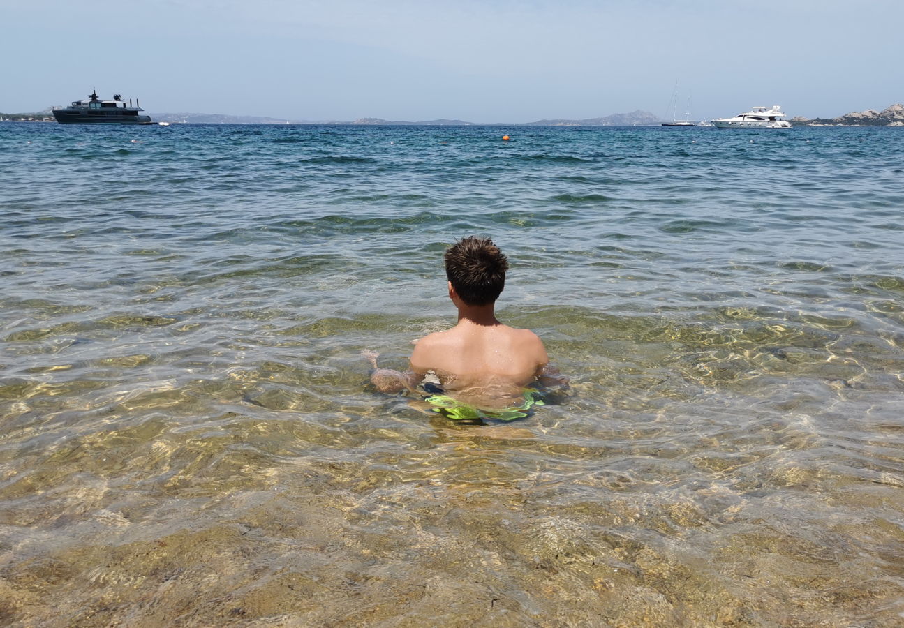 Ein Junge sitzt im glasklaren Wasser des Mittelmeeres und beobachtet Motoryachten.