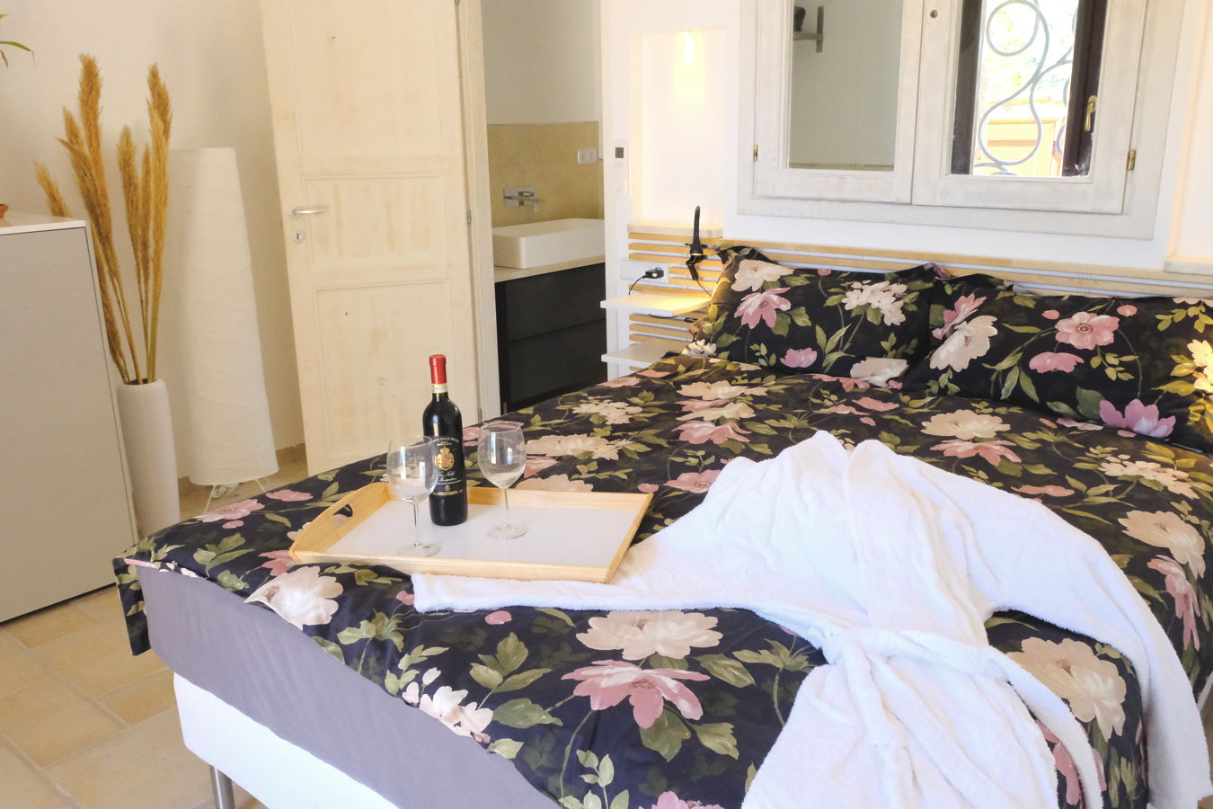 Bett mit bunter Bettwäsche, auf dem ein weißer Bademantel liegt und ein Tablett mit einer Flasche Rotwein und zwei Gläsern.
